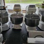 bakkerij machine kopen - Een rij bakkerijmachines, waaronder industriële deegmixers van verschillende merken, gepositioneerd op een vloer met een raam op de - theovanvliet.nl